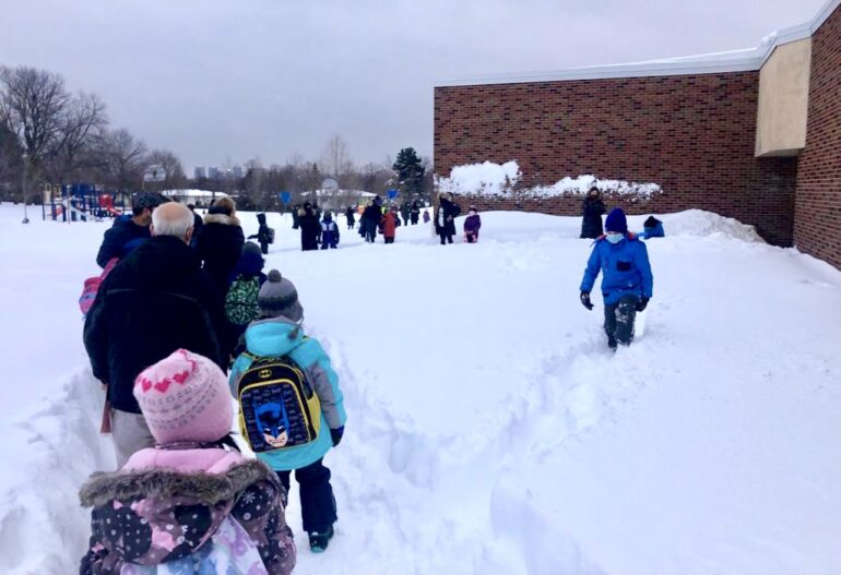 Parents and children line up to enter school on snowy morning.