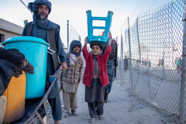 A displaced family in Kandahar. A man in Afghan clothes is pushing a wheelbarrow with two tubs in it. One blue and one orange. Behind him are two young boys. One is holding a blue stool on his head. They are standing besides a chain link fence with people beyond it. Behind them is a man in camouflage and holding an assault rifle