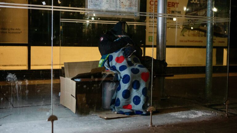 A homeless man sleeps in a bus shelter, in Toronto, on March 11, 2022. The city announced the hiring of 15 new outreach workers to help homeless people.