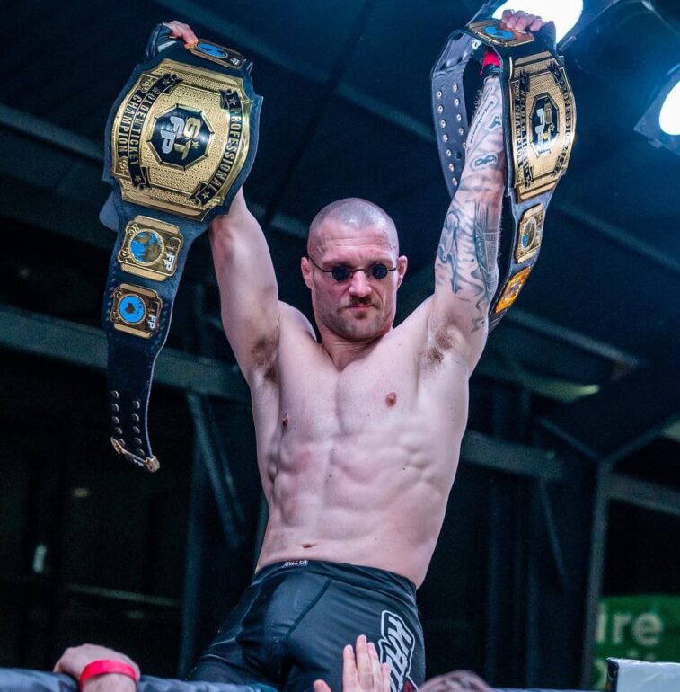 Thomas Paull sits atop cage raising both championship belts following his first round knockout victory over Perry Goodwin at GTFP 18.