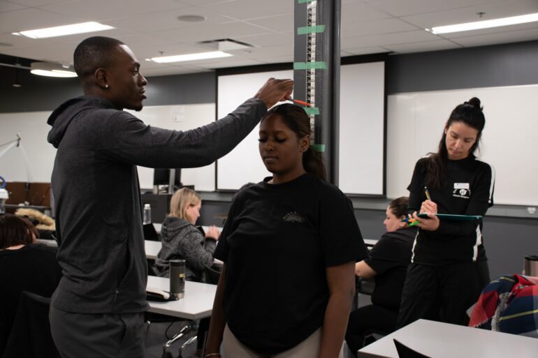 From left to right, ESLM student Tarik Smith-James is taking his client's height as a part of his client assessment as an ESLM professional. His colleague, Alejandra Ramirez Ovalle, is writing the measurements down and preparing the next step in their client assessment, which is the client's weight documentation.