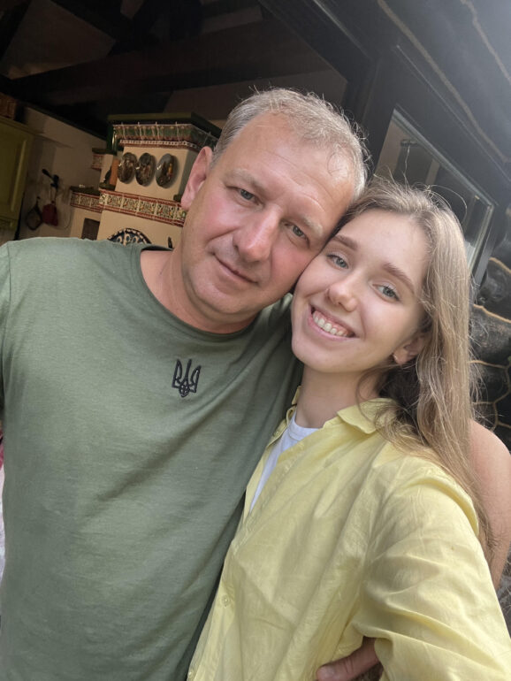 A middle-aged man wearing a green shirt posing with his daughter, a blond haired girl in a yellow shirt.