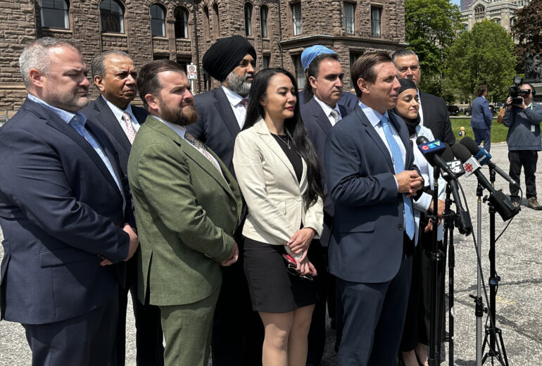 A group of people stand in front of a microphone outside of Queen's Park. Brampton Mayor Patrick Brown is directly in front of the main microphone speaking.