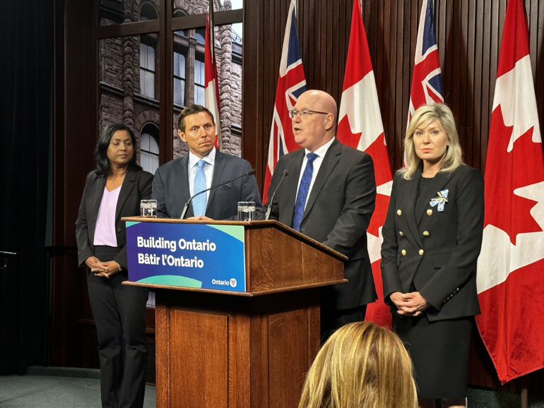 Municipal Affairs and Housing Minister Steve Clark stands in front of a podium at Queen's Park. The mayors from Brampton, Mississauga and Caledon stand beside him.