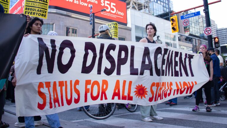 Three people hold a banner reading "NO DISPLACEMENT, STATUS FOR ALL, STATUS NOW"
