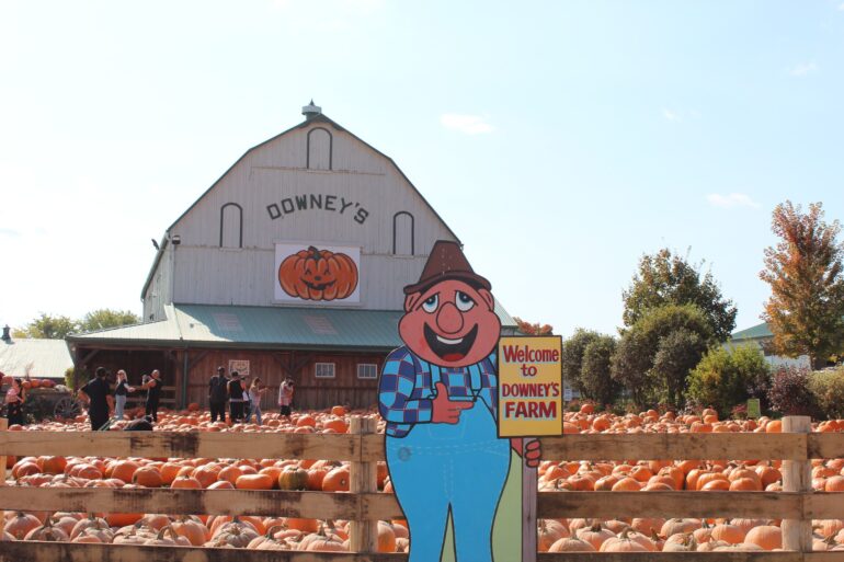 A stand-up board of a farmer holding a welcome sign and in the background there is a pumpkin patch and farm house that says "Downey’s".