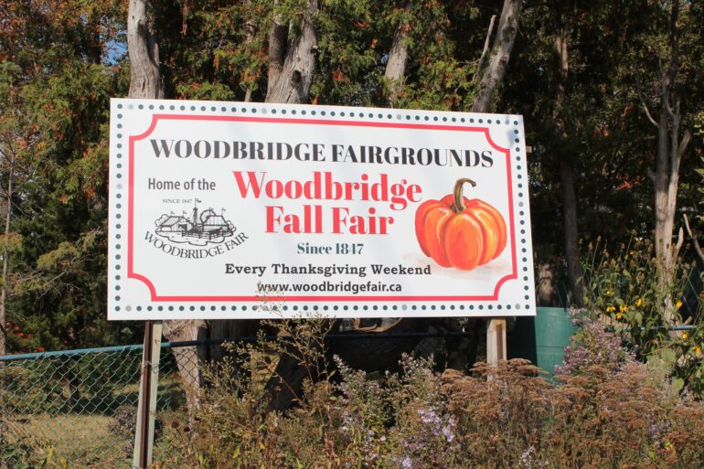 A white sign that says "Woodbridge Fairgrounds" in black coloured text and "Woodbridge Fall Fair" in a bright red colour.
