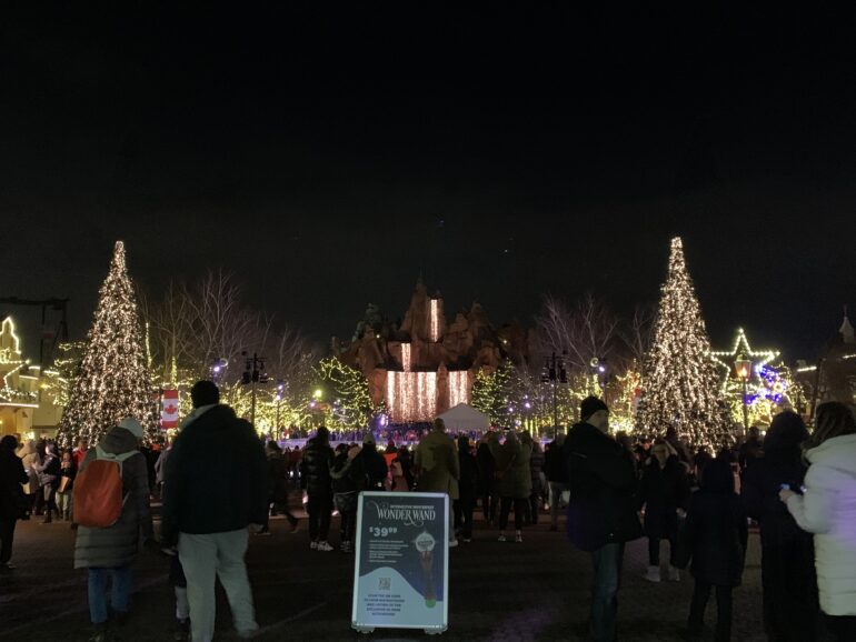 Groups of people at the front of Canada' Wonderland Winterfest. In the background there is mountain in the middle and two Christmas trees on both sides.
