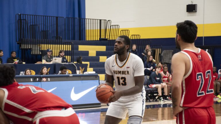 Humber basketball player Jamani Barrett, wearing a white basketball jersey wearing number 13. He is at the free throw line about to shoot the ball in the basketball net.