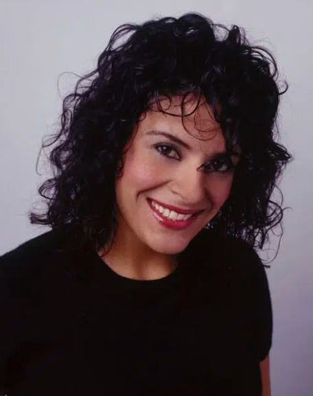 A person with mid-length curly hair in a black T-shirt in front of a white background.