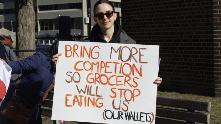 A member of ACORN, Olivia Baker holding a placard 'Bring more competition so grocers will stop eating us {our wallets)'