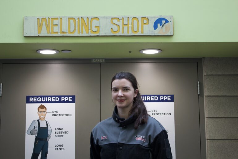 A girl in her welding uniform is smiling in front of the entrance of the welding lab.