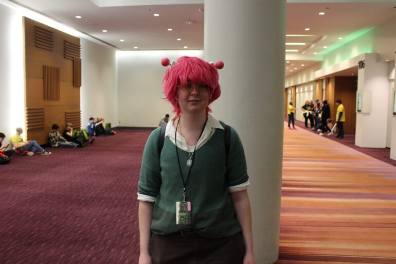 A person standing in a hall, dressed in a green shirt with a white collared shirt underneath and brown pants. Wearing a pink wig with pink bulbed antenna on both sides of their head.