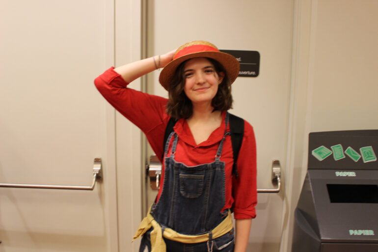 A girl dressed in a red long sleeve blouse, with dark blue jean overalls, and a yellow waist tie. She is also wearing a straw hat with a red accent while smiling at the camera.