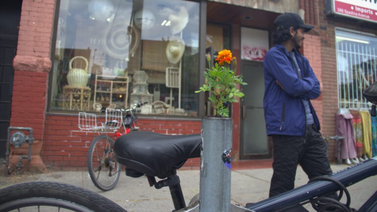 A flower sapling planted in a bike ring post with a man standing in the background.